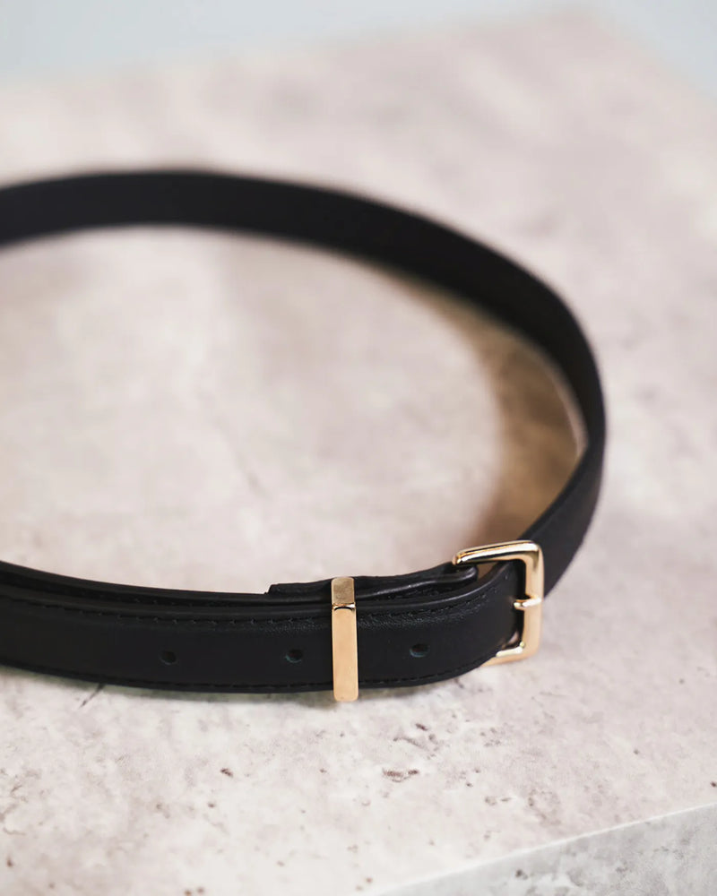 Black leather belt with gold buckle and keeper coiled on a marble surface. The Trouser Belt Gold Black.