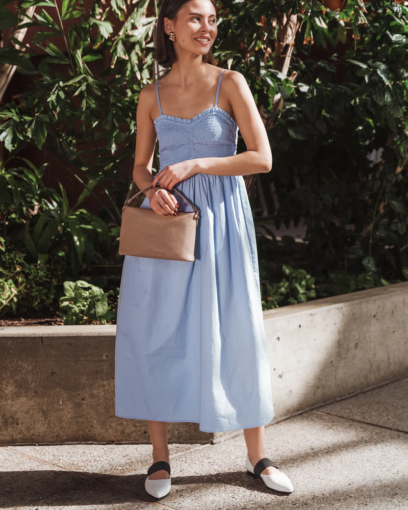 Woman in a blue sundress carrying a pebbled leather mini bag with top handle. The Via Monte Bag Biscotti.