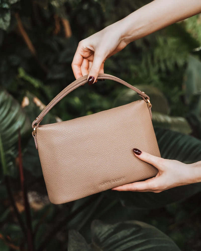 Hands holding a pebbled leather mini bag with top handle against tropical greenery. The Via Monte Bag Biscotti.