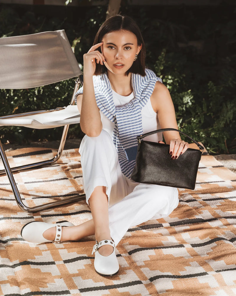 Woman seated on a rug outdoors carrying a pebbled leather mini bag with top handle. The Via Monte Bag Black.