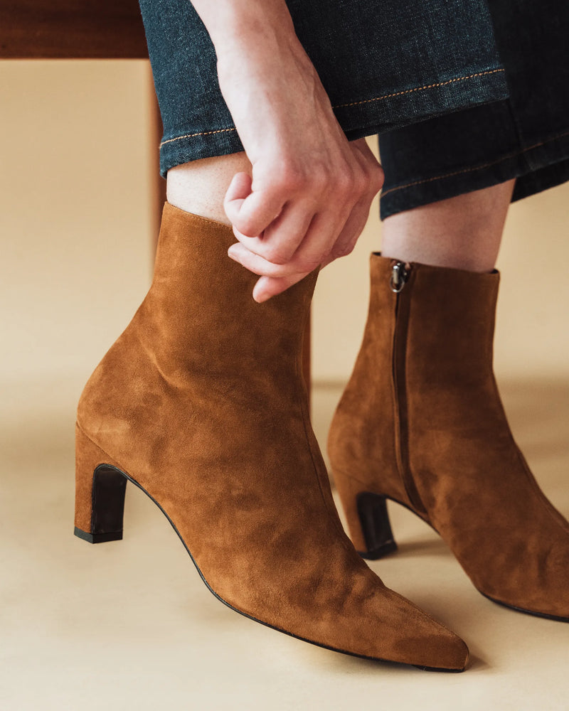 Woman seated in dark denim jeans wearing The Toujours Boot Cinnamon, hand resting on ankle. Sizes 5–12, wide available.
