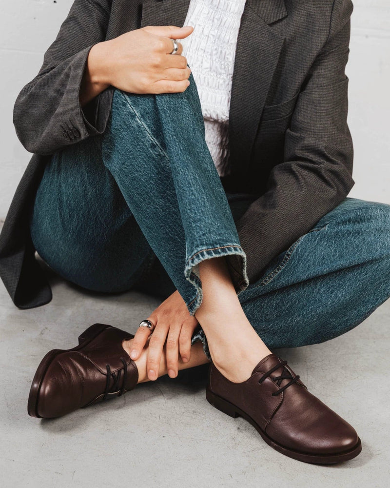 Model seated wearing the leather oxfords with denim. The Capsule Oxford Cocoa Plum. Sizes 5–12, wide available.