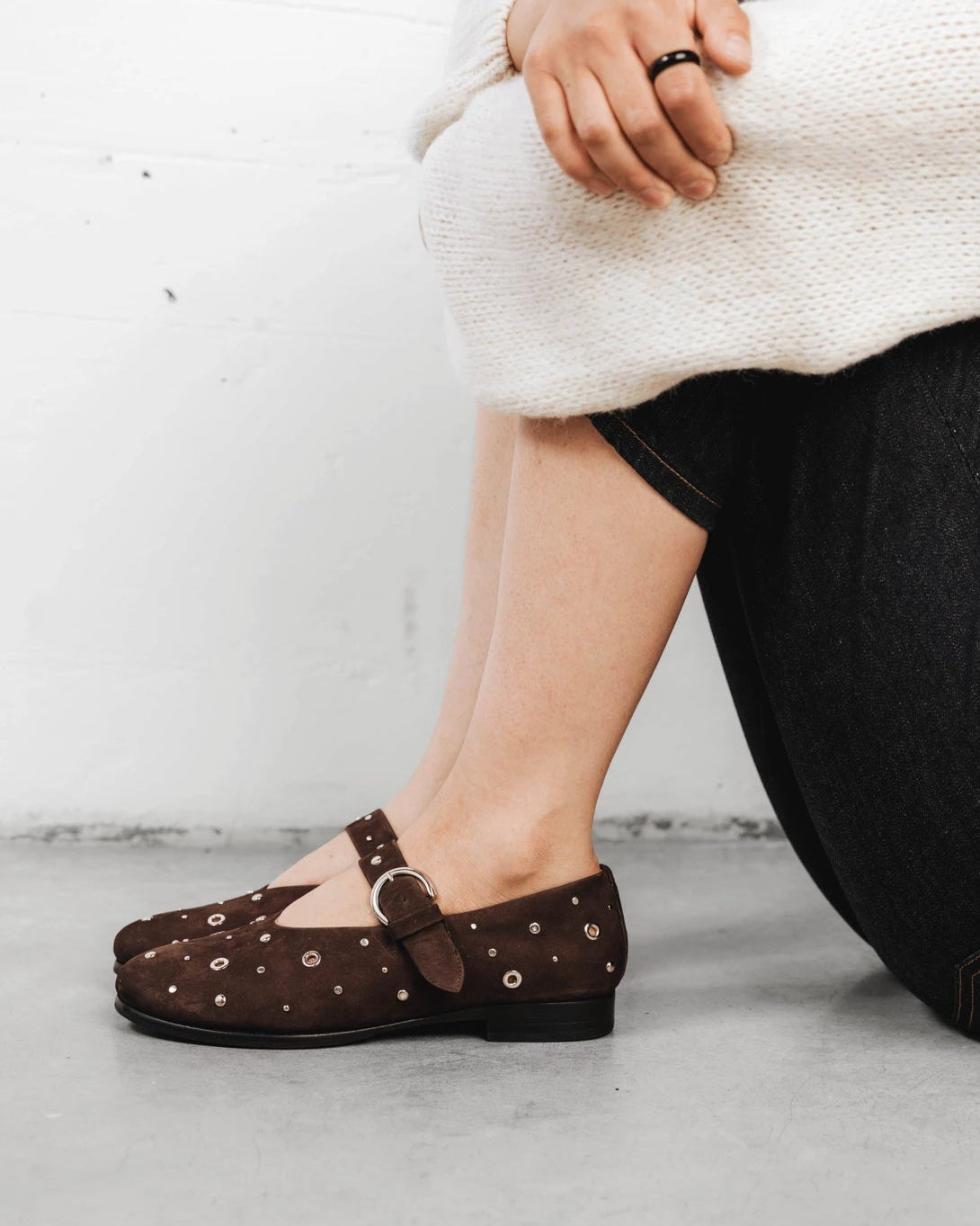 Person kneeling wearing studded suede mary janes. The BLVD Mary Jane Cocoa Plum Studded. Sizes 5–12, wide available.