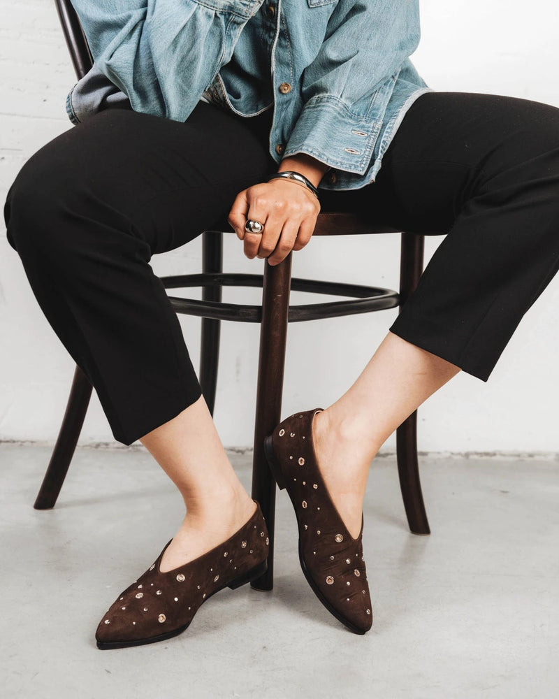 Model seated wearing the studded suede flats. The Atelier Flat Cocoa Plum Studded. Sizes 5–12, wide available.