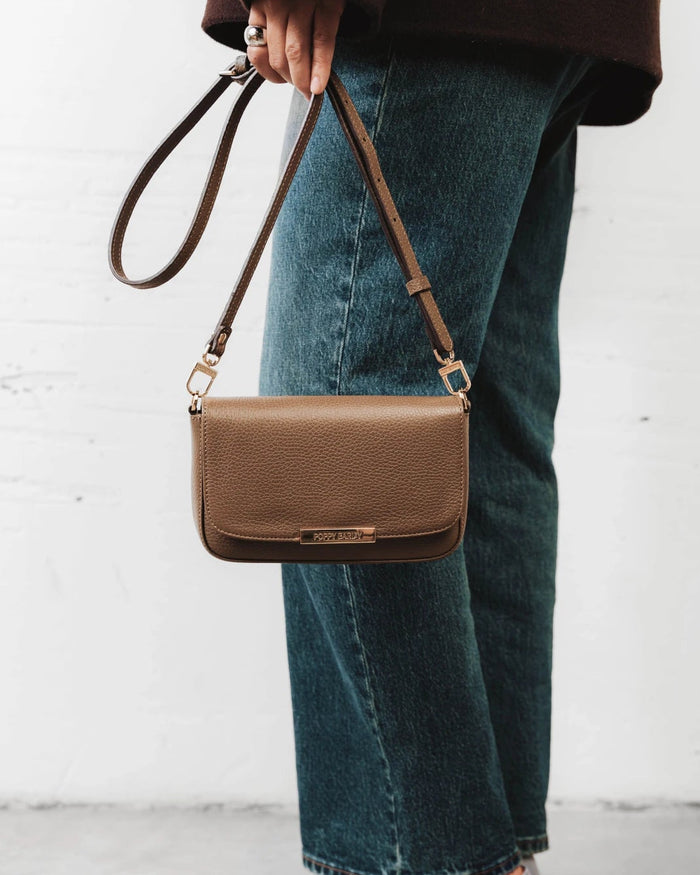 Front view of the leather flap bag with a long adjustable strap laid flat. The Day To Night Bag Tapenade.